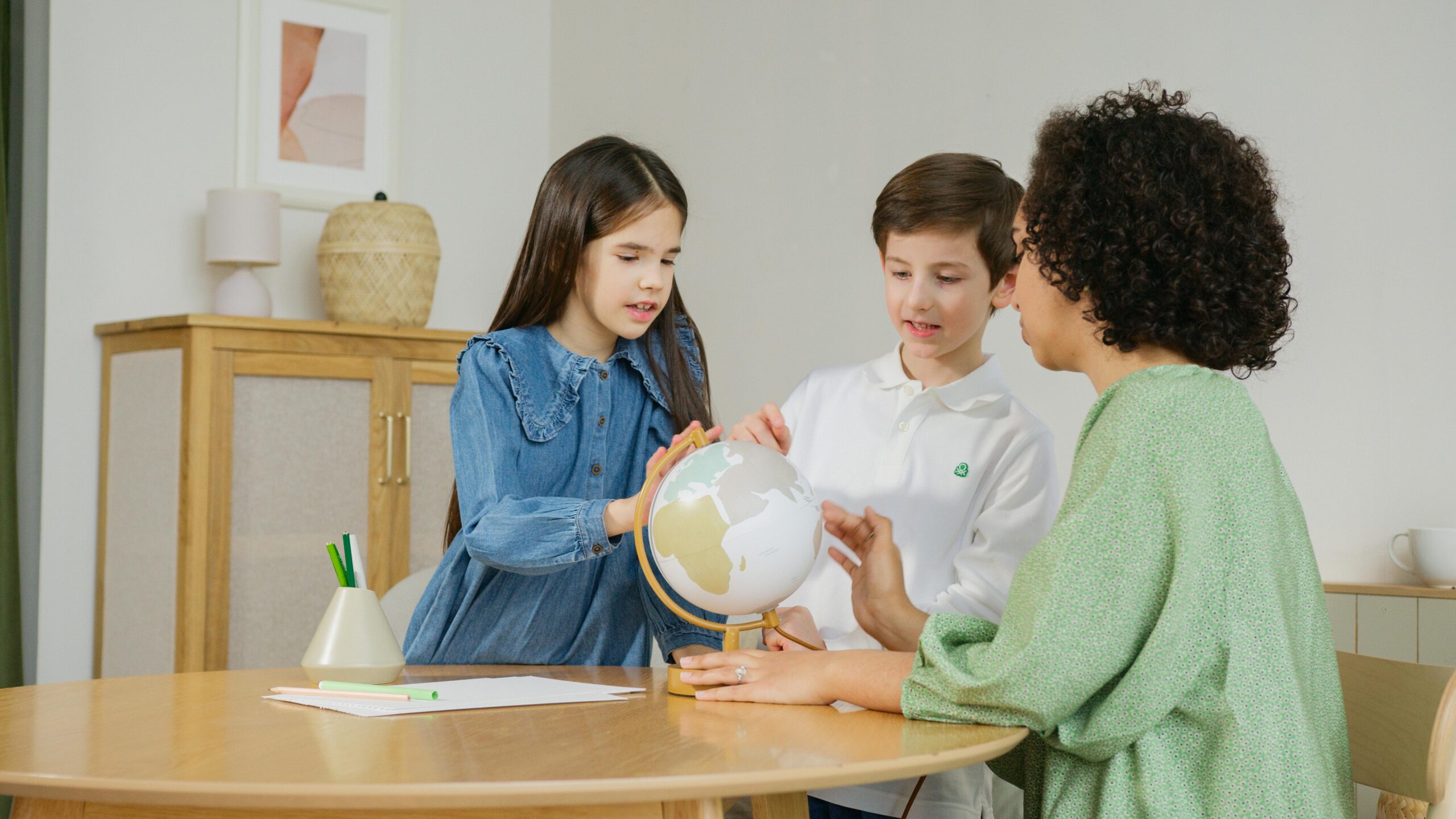 A woman and two children engage in an educational activity at a round table, exploring a globe in a cozy, modern setting.