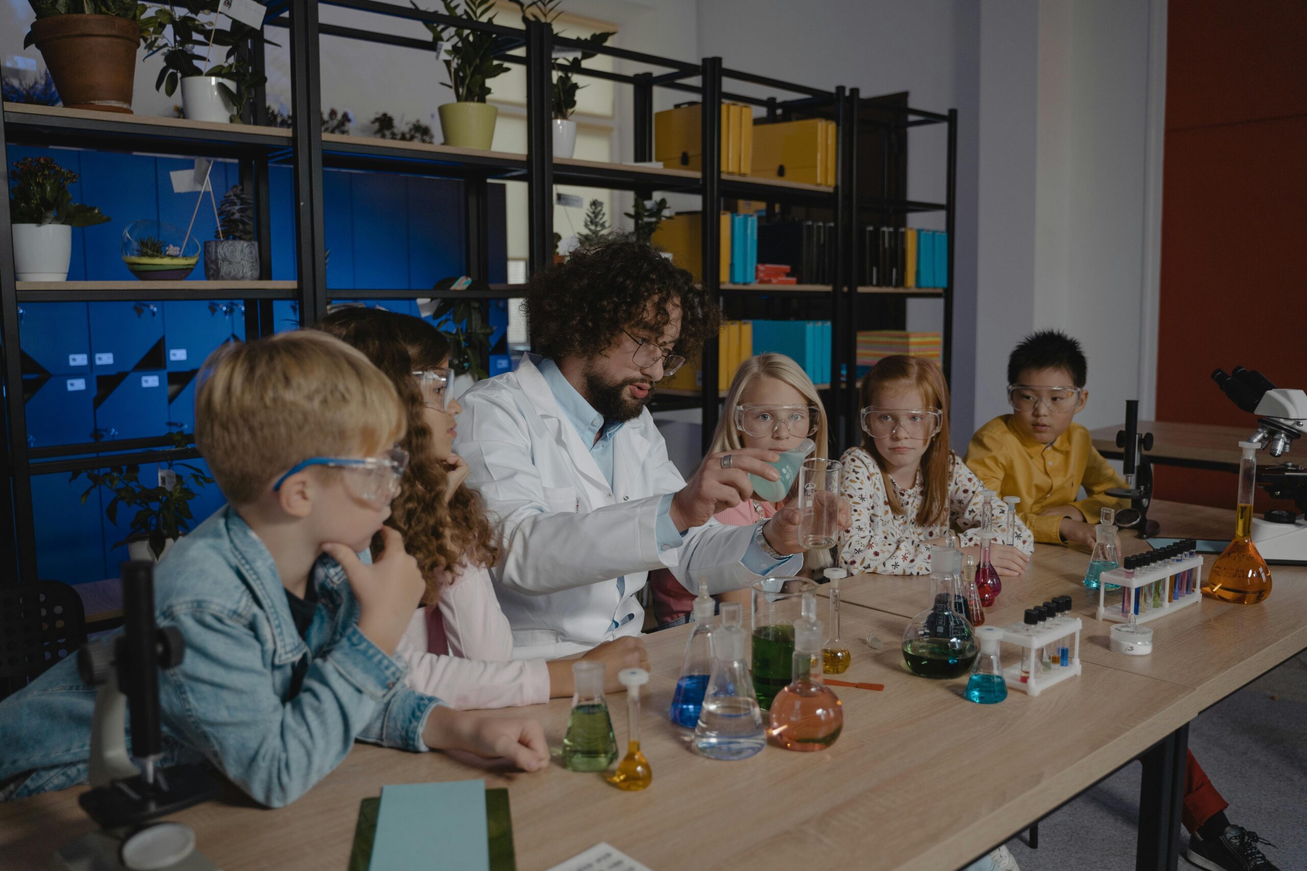 A teacher in a lab coat demonstrates a science experiment to a group of children, surrounded by colorful glass beakers and plants.