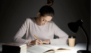 A person in a light sweater writes in a notebook at a desk, illuminated by a lamp, with coffee and books nearby.