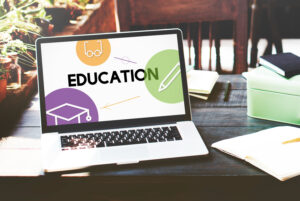 A laptop displaying the word EDUCATION alongside educational icons, set on a wooden desk with notebooks and stationery.