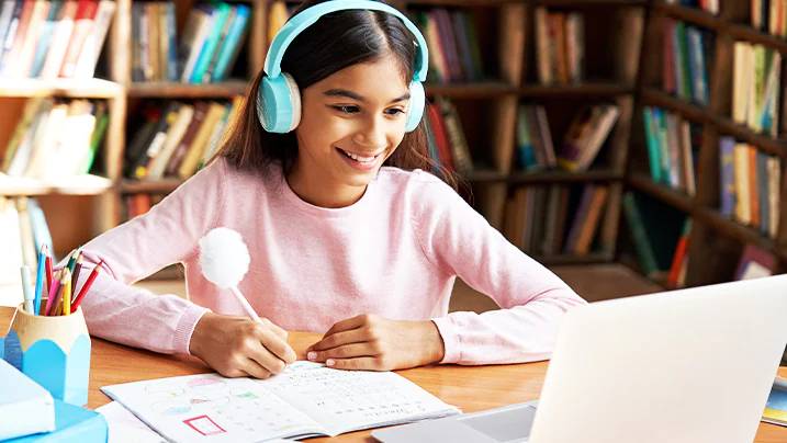A student wearing teal headphones sits at a desk, writing in a notebook with a laptop open and bookshelves in the background.