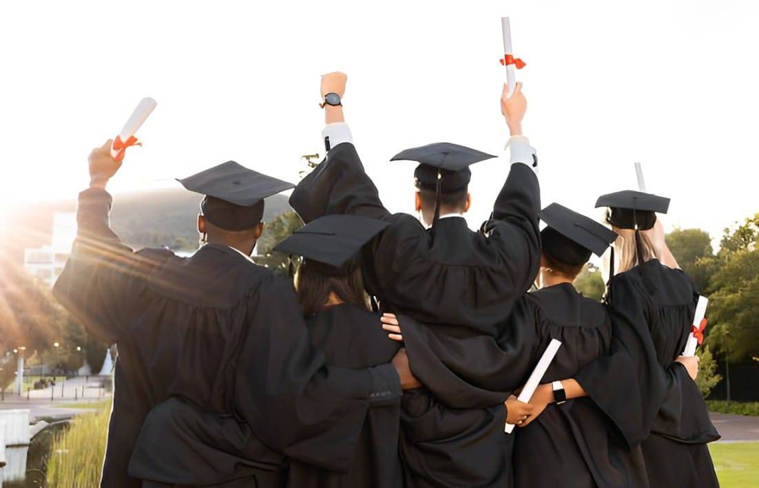 A group of six graduates in black gowns and caps joyfully raise their diplomas against a sunset backdrop, celebrating their achievement.