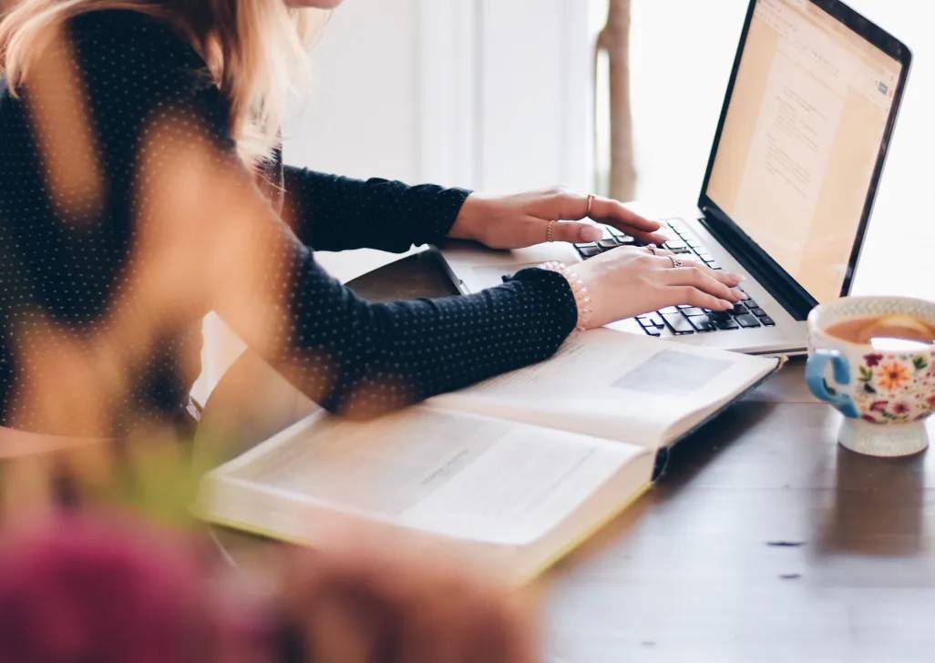 A woman types on a laptop at a wooden desk, surrounded by an open book and a colorful teacup, with soft natural light illuminating the scene.