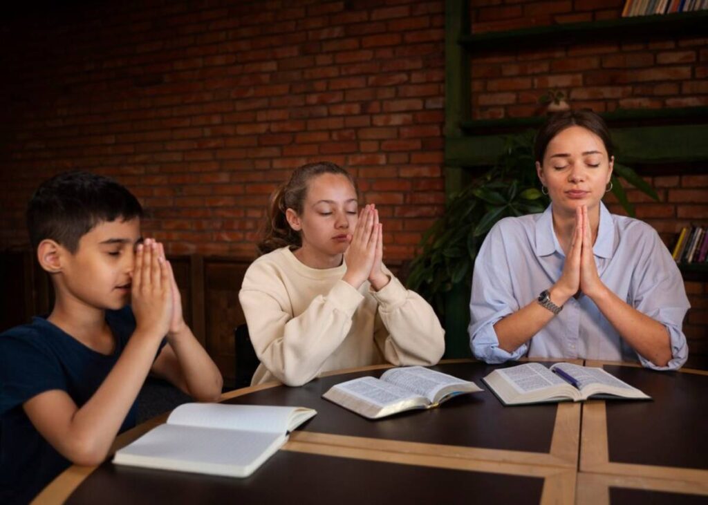 Three people seated at a table with open books, with hands clasped in prayer, against a brick wall and plant backdrop.