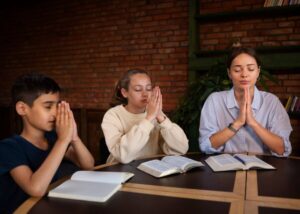Three people seated at a table with open books, with hands clasped in prayer, against a brick wall and plant backdrop.