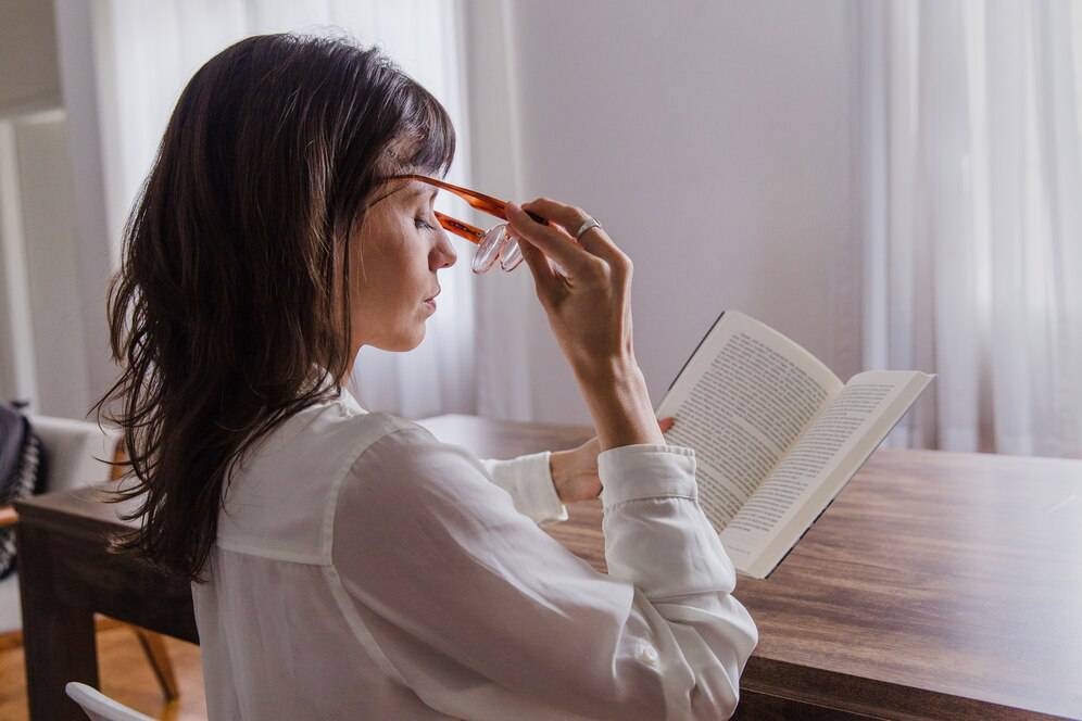 A person in a white shirt sits at a wooden table, reading a book and thoughtfully adjusting their glasses in a bright, airy room.