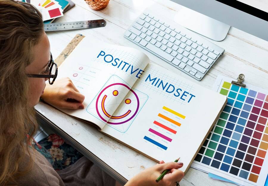 A person sketches in a notebook showing the phrase Positive Mindset, next to a colorful palette and a computer keyboard.