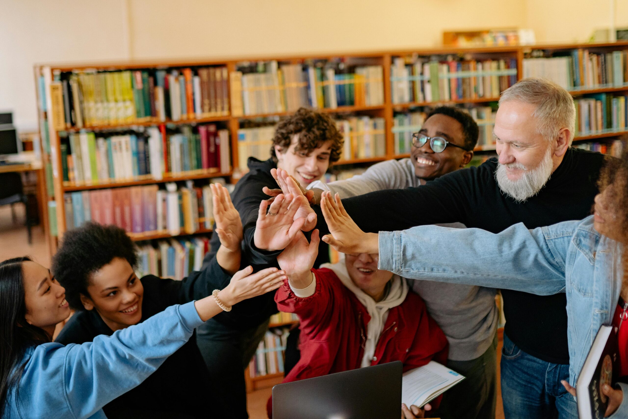 A group of diverse individuals enthusiastically reaching out to each other in a library setting, surrounded by bookshelves.