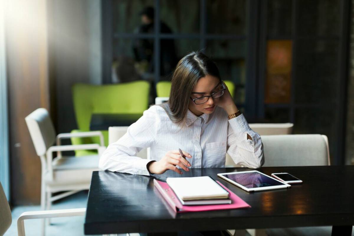 A person sits at a table in a café, writing notes on a notepad, with a tablet and smartphone beside them.