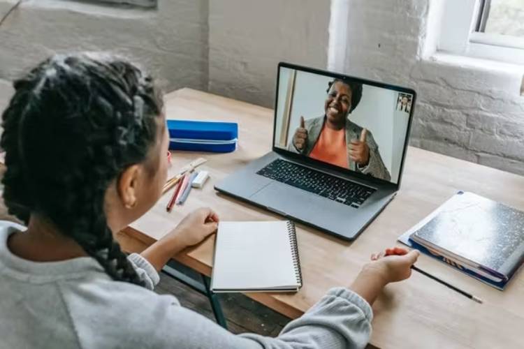 A young person with braided hair sits at a desk, engaged in a video call on a laptop, with a notebook and stationery visible.