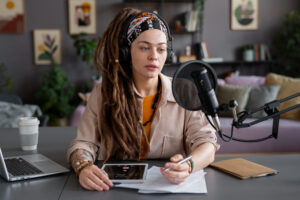 A person with long dreadlocks wears headphones and holds a pen, sitting at a desk with a laptop, tablet, and coffee cup, ready to record.