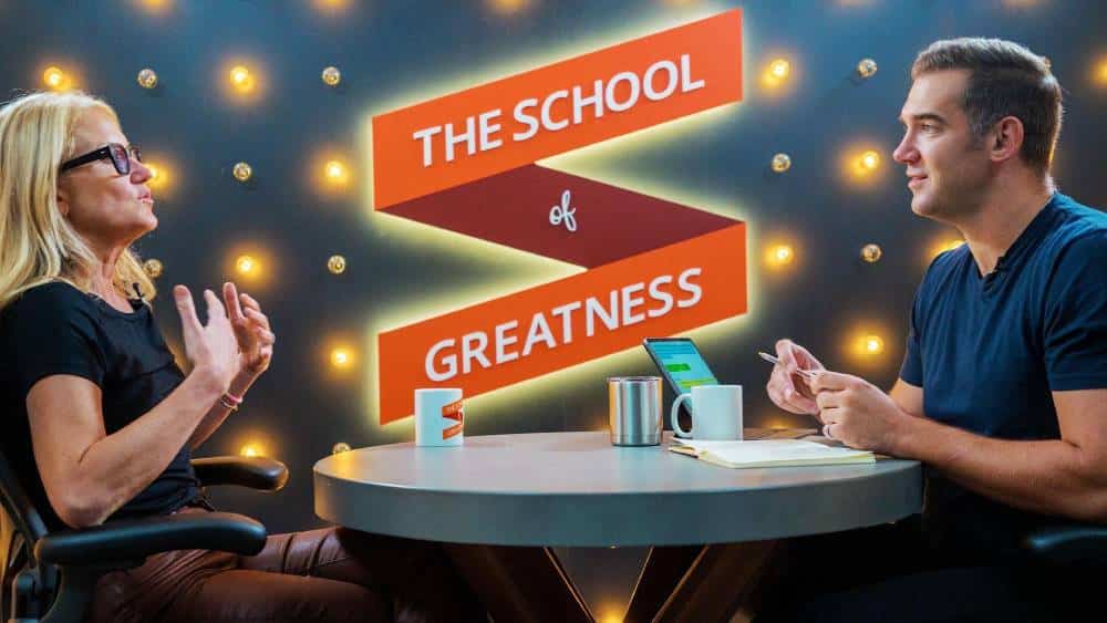 Two people engage in a discussion at a round table, with a backdrop featuring the text THE SCHOOL of GREATNESS and bright lights.
