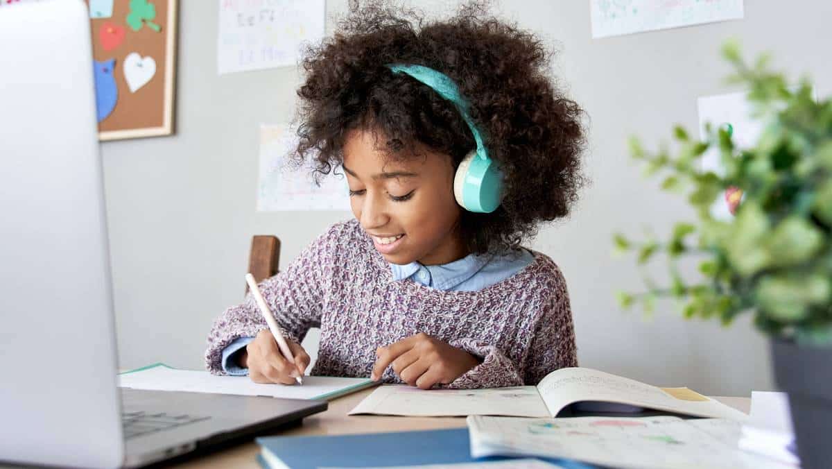 A child wearing headphones is focused on writing at a desk, surrounded by workbooks and colorful drawings on the wall.