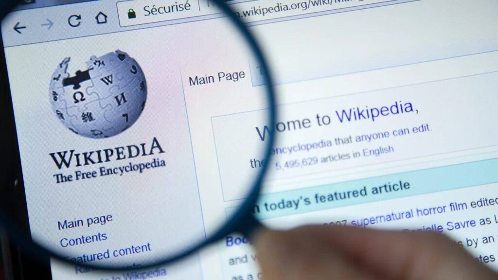 A close-up of a computer screen displaying the Wikipedia homepage, with a magnifying glass highlighting the logo and main page link.