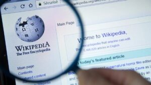 A close-up of a computer screen displaying the Wikipedia homepage, with a magnifying glass highlighting the logo and main page link.