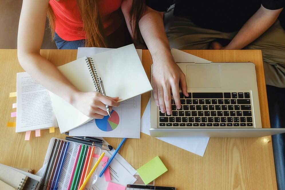 A person's hand writes in a notebook while another hand types on a laptop, surrounded by colorful stationery on a wooden table.