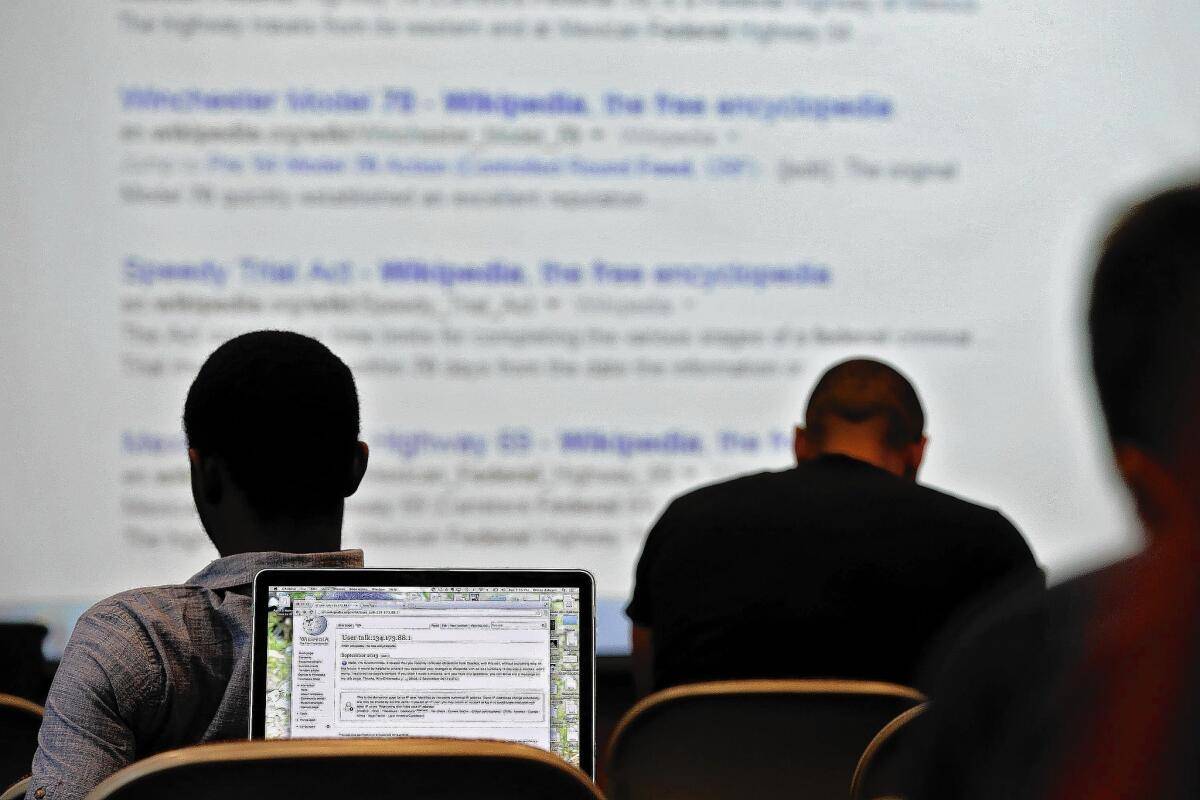 Two people are seated with their backs to the camera; one has a laptop open, displaying a Wikipedia page, while the other views a projector screen.