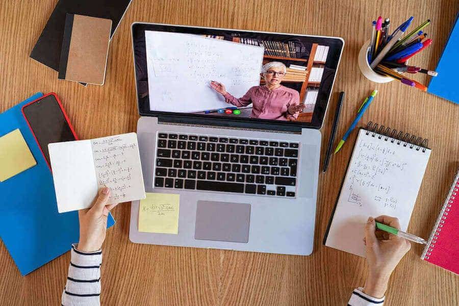 A laptop displays an online lesson with a whiteboard, surrounded by notebooks, sticky notes, and writing tools on a wooden table.