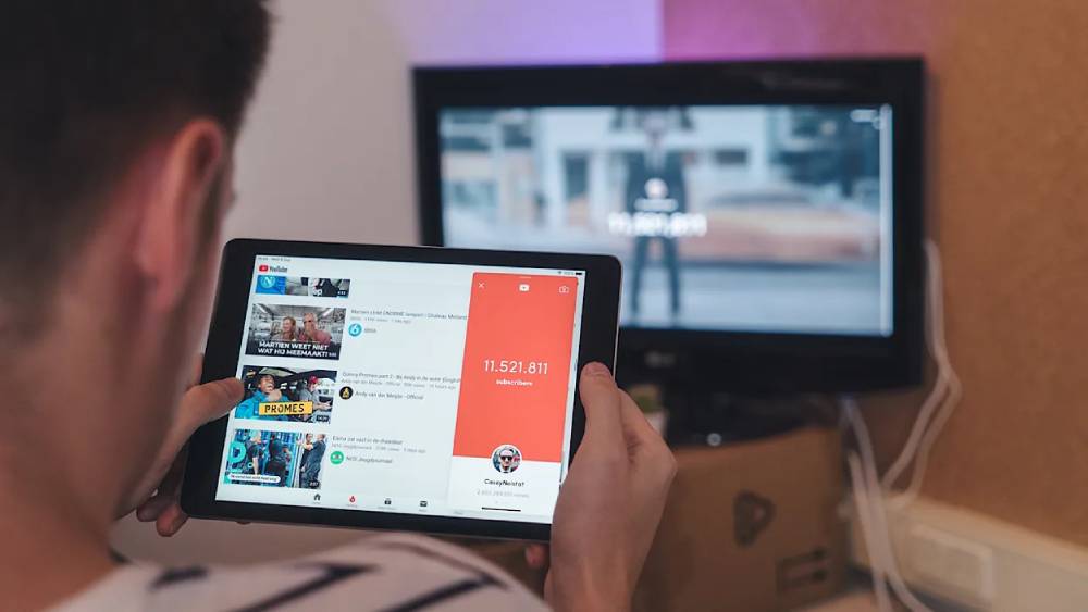 A person watches YouTube on a tablet, displaying videos and subscriber count, with a television screen showing another video in the background.