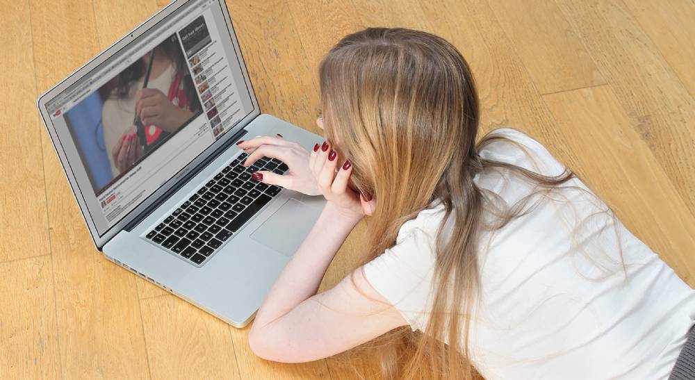 A young woman with long hair lies on the floor, focused on a laptop screen showing a YouTube video.