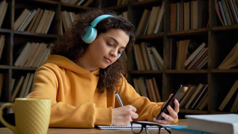 A person in a yellow hoodie wears headphones, writing notes while holding a smartphone, with a bookshelf in the background.