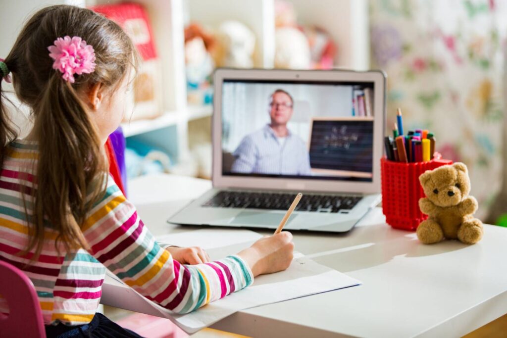 A girl writes in a notebook while coding with a laptop displaying an online class, next to colorful pencils and a teddy bear.
