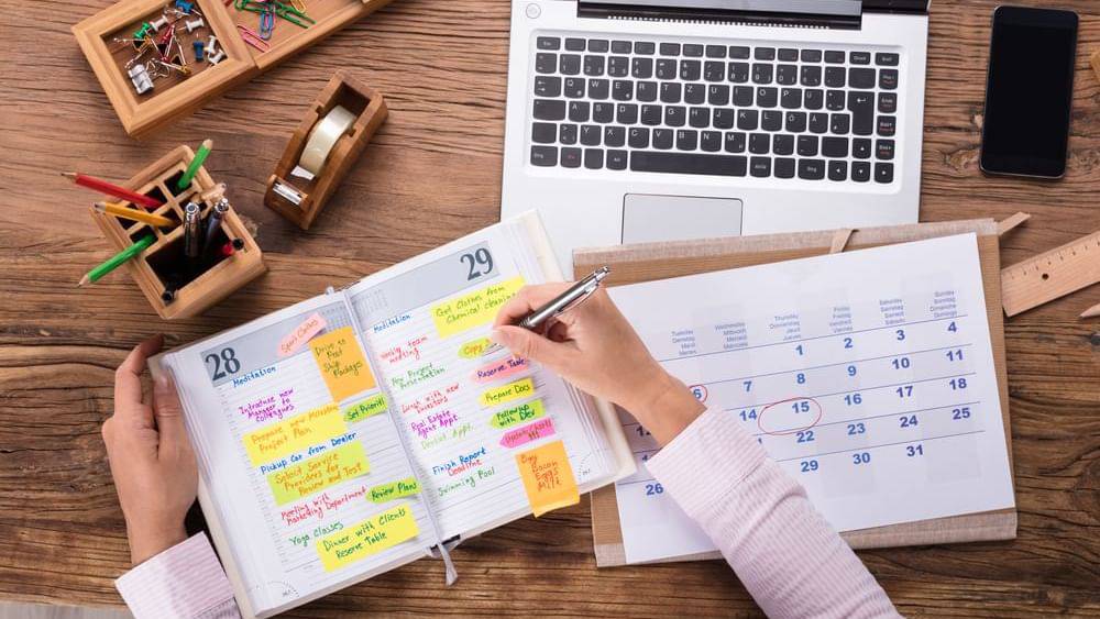 A person writing in a planner filled with colorful notes, surrounded by a laptop, calendar, and stationery on a wooden desk.