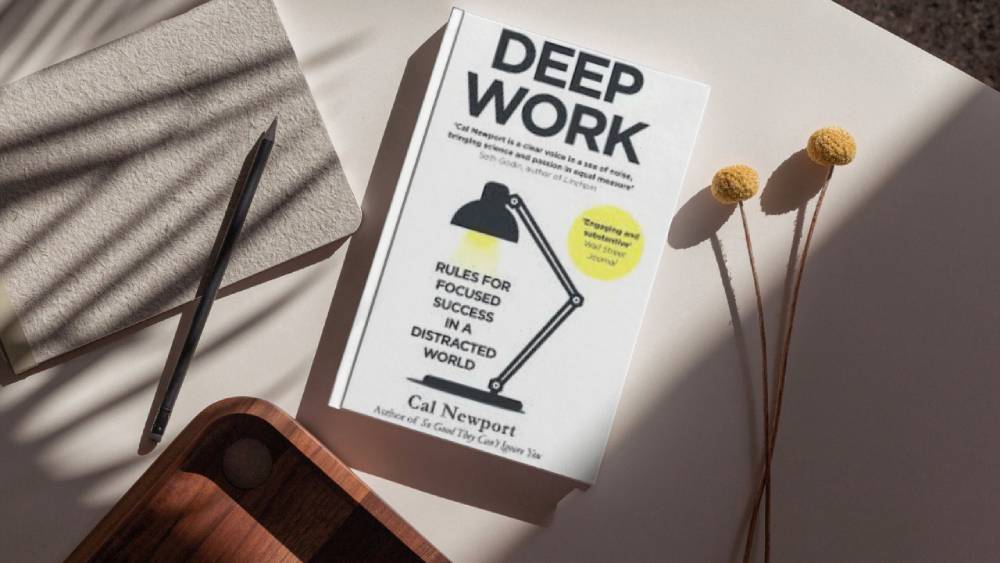 A flat lay featuring Deep Work by Cal Newport, a notebook, pen, and decorative flowers atop a light surface.