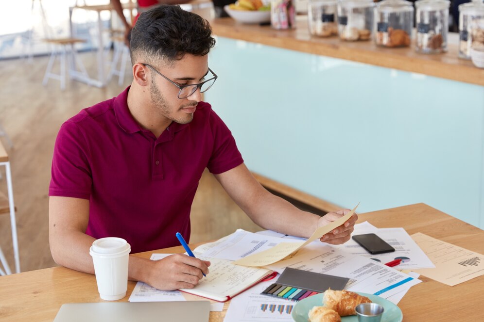 A person in a maroon shirt sits at a wooden table, reviewing documents with coffee, pastries, and stationery scattered around.