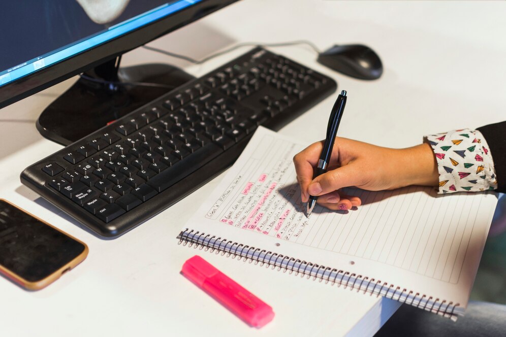 AA hand writes notes in a notebook, with a computer screen, keyboard, and smartphone visible on the desk, alongside a highlighter.