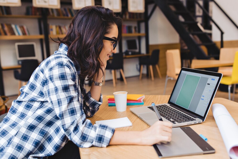 A person in a plaid shirt sits at a desk, focused on a laptop, with a notebook and coffee cup nearby in a cozy study environment.