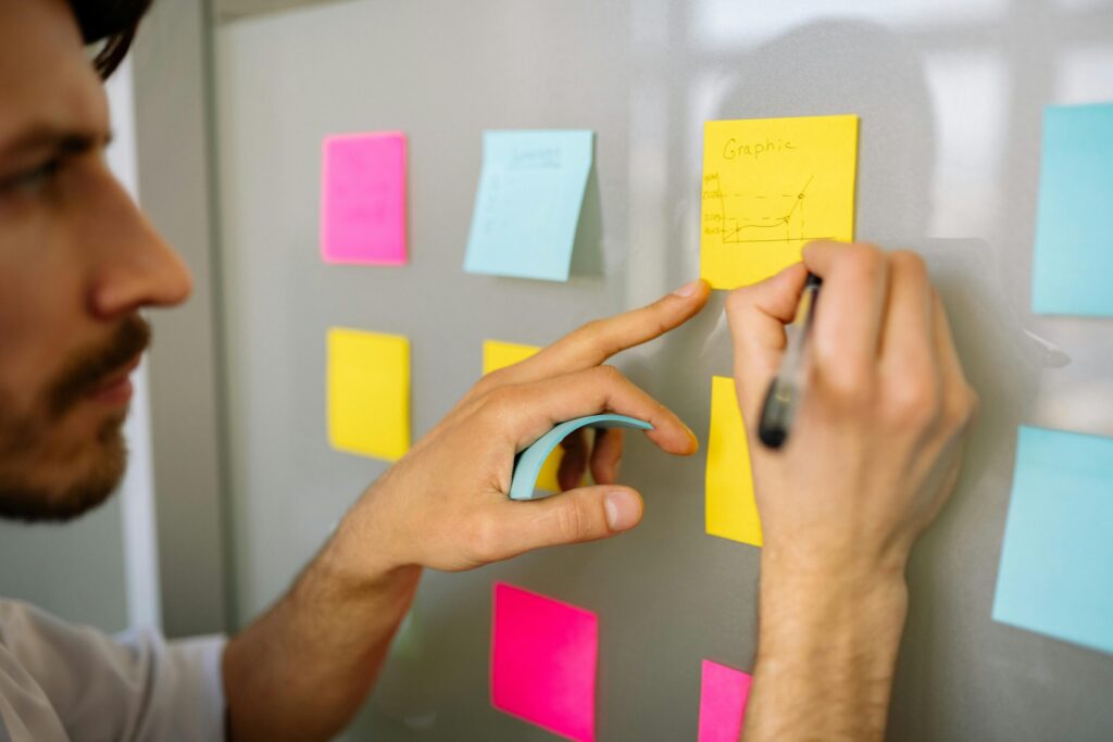 A close-up of a hand writing on a yellow sticky note labeled Graphic while placing it on a board surrounded by various colored notes.