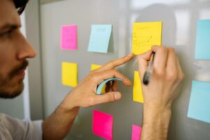 A close-up of a hand writing on a yellow sticky note labeled Graphic while placing it on a board surrounded by various colored notes.