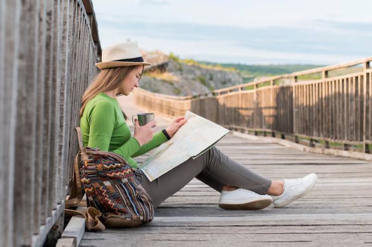 A person sits on a wooden walkway, holding a map and a mug, wearing a green sweater and a hat, with a colorful backpack beside them.
