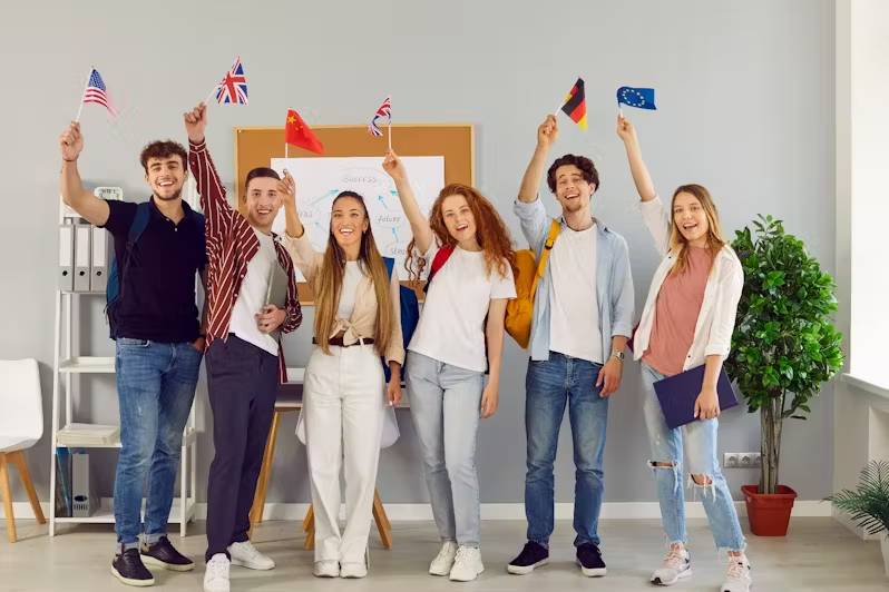 A diverse group of students joyfully holds small international flags, standing together in a modern classroom with greenery in the background.