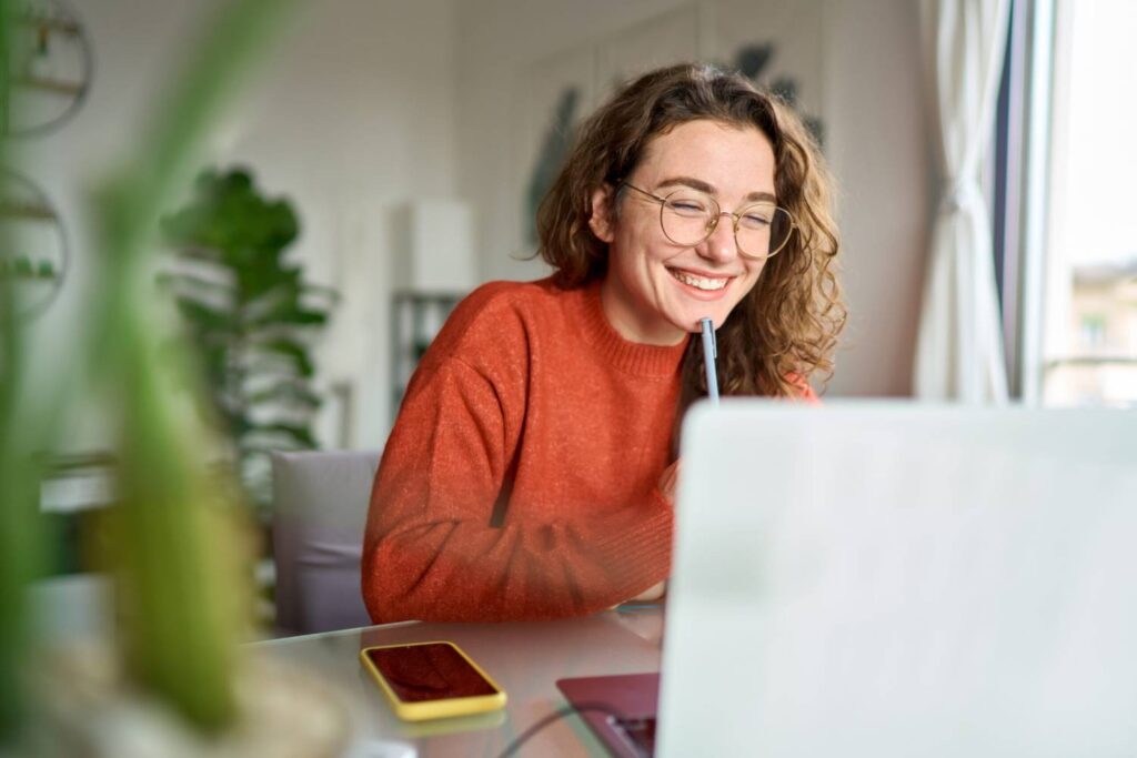 A person in an orange sweater sits at a desk with a laptop and smartphone, surrounded by indoor plants and natural light.
