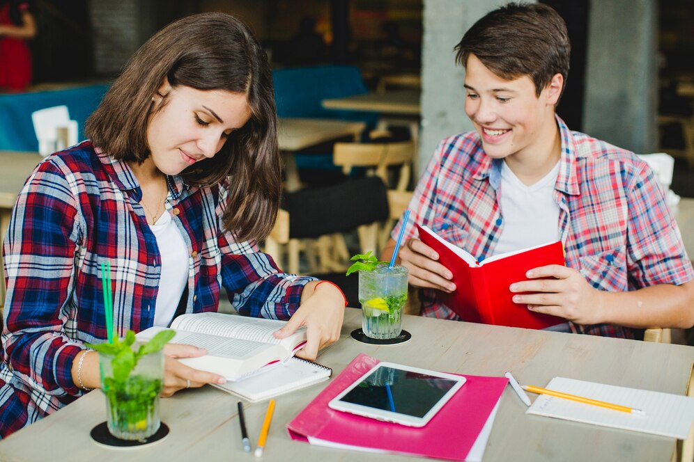 A young woman reads a book, while a young man holds a red book, both seated at a table with drinks and study materials around them.