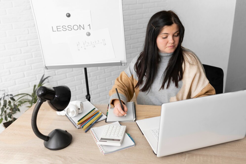 A person is seated at a desk, taking notes with a pen, surrounded by stationery, a laptop, and a whiteboard displaying LESSON 1.