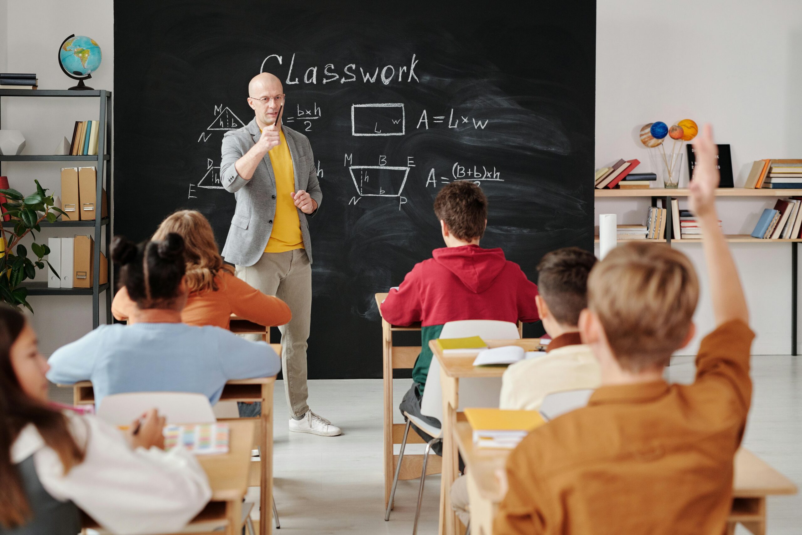 A teacher stands at a blackboard with mathematical equations while students raise their hands, engaged in classwork.