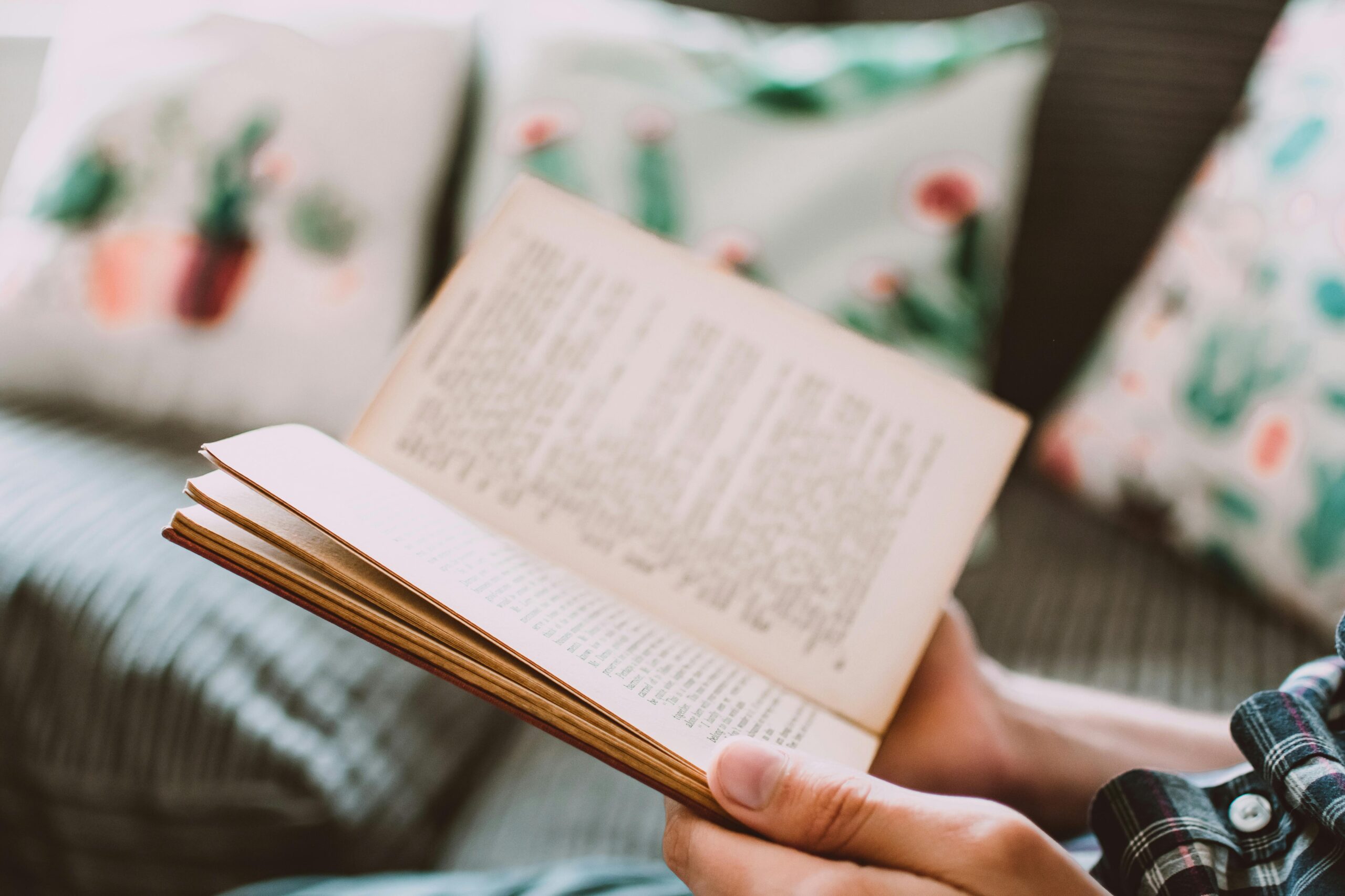 A person holds an open book, revealing pages filled with text, while sitting on a couch adorned with floral-patterned pillows.