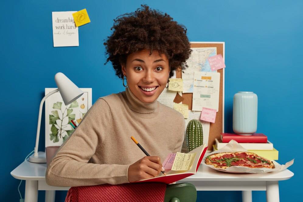 A person sitting at a desk, writing in a notebook with colorful sticky notes, surrounded by a lamp, a plate of pizza, and a corkboard.