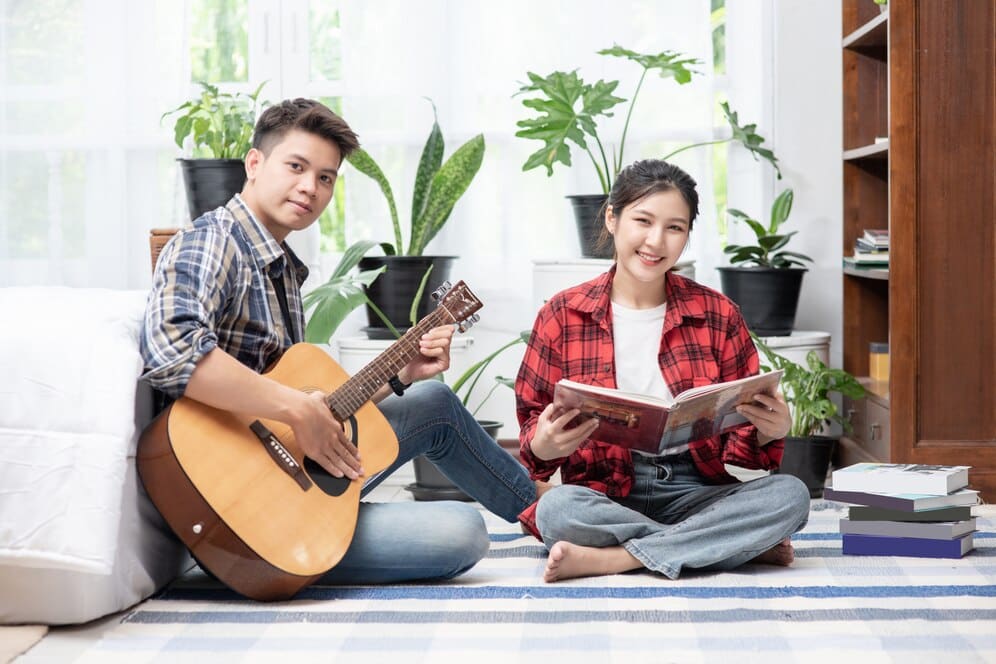 A person sits with a guitar while another reads a book, surrounded by houseplants and books on a cozy carpeted floor.