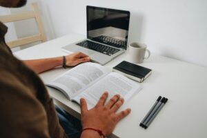 Person reading a book on a white desk with a laptop, a coffee mug, a notebook, and three pens nearby.