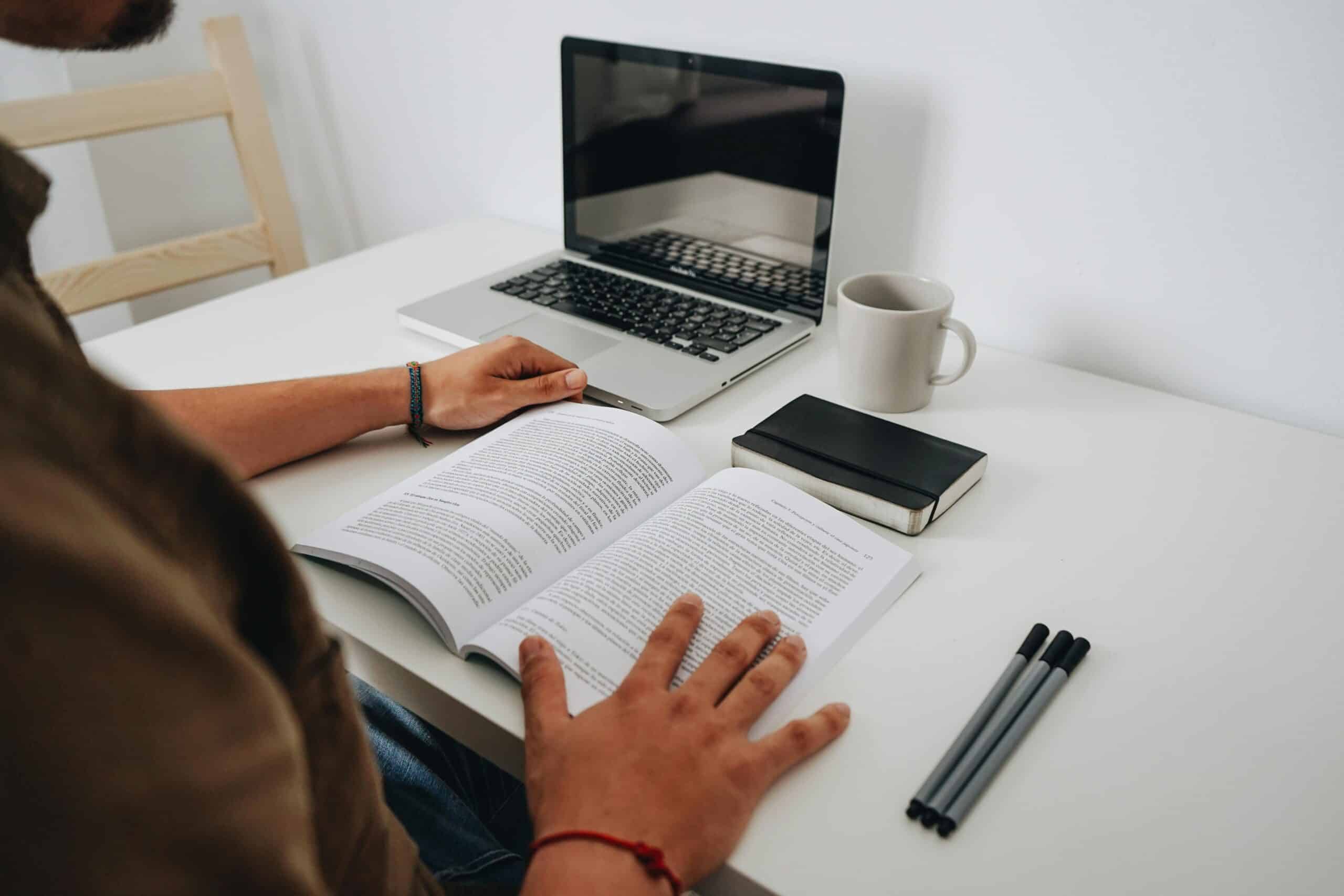 Person reading a book on a white desk with a laptop, a coffee mug, a notebook, and three pens nearby.