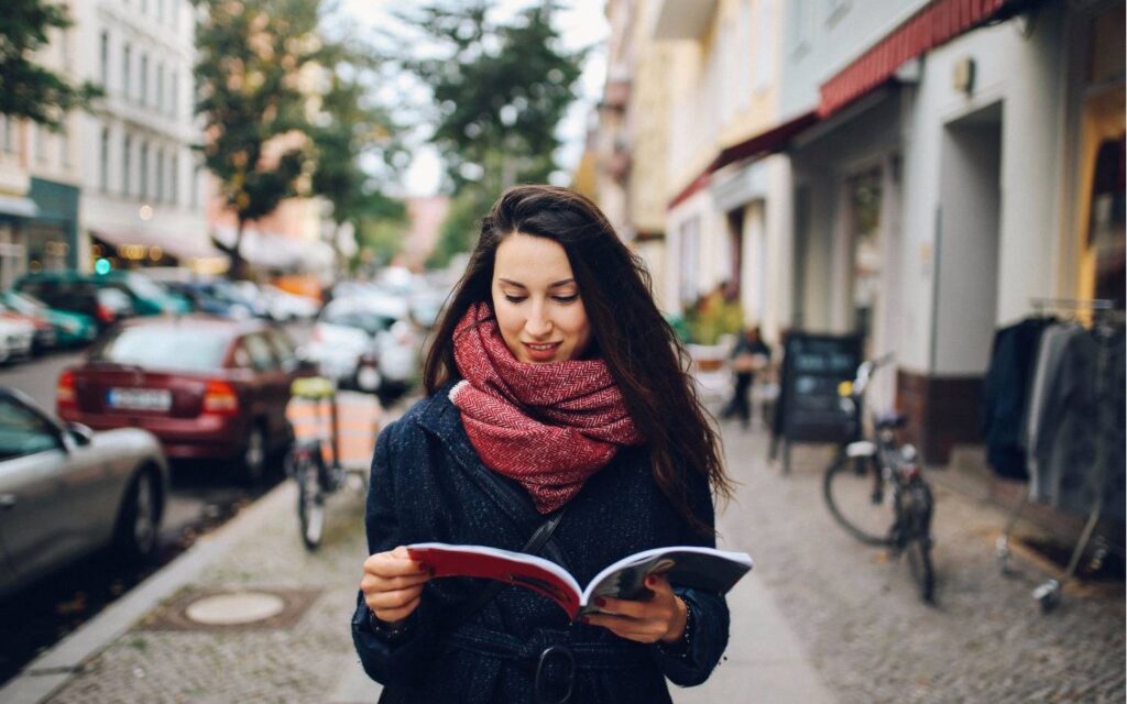 A person stands on a bustling street, reading a magazine while dressed warmly in a dark coat and a patterned scarf. Cars and bikes are nearby.