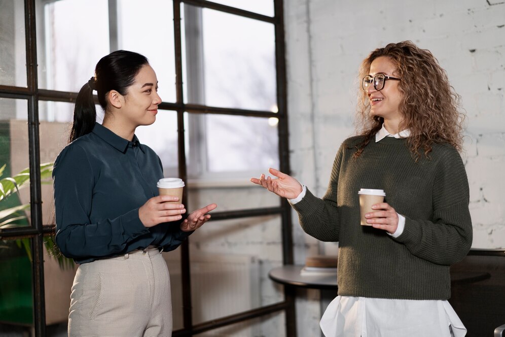 Two women engage in conversation while holding coffee cups in a modern workspace with large windows and a minimalist aesthetic.