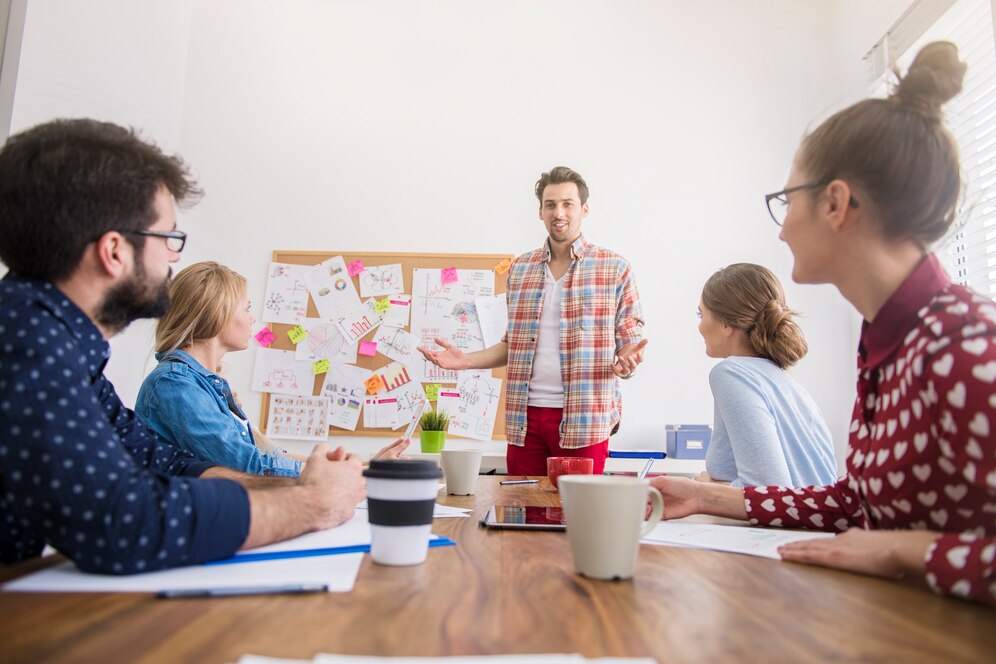 A group of professionals engages in a meeting, discussing ideas and projects while a presenter gestures towards a board covered in notes.