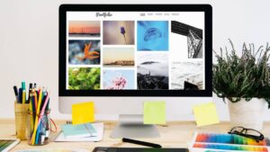 A computer screen displays a photo portfolio with colorful images surrounded by office supplies and a potted plant on a wooden desk.