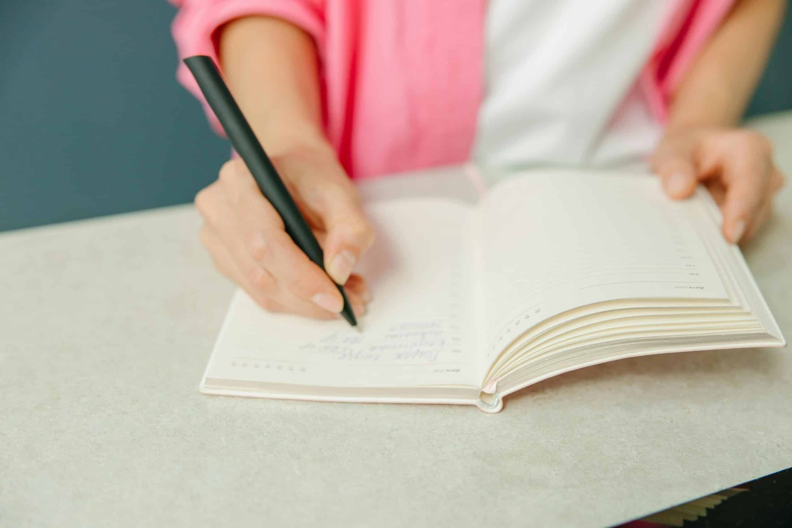 A close-up of a person's hand holding a black pen, writing in a blank notebook on a light gray table.