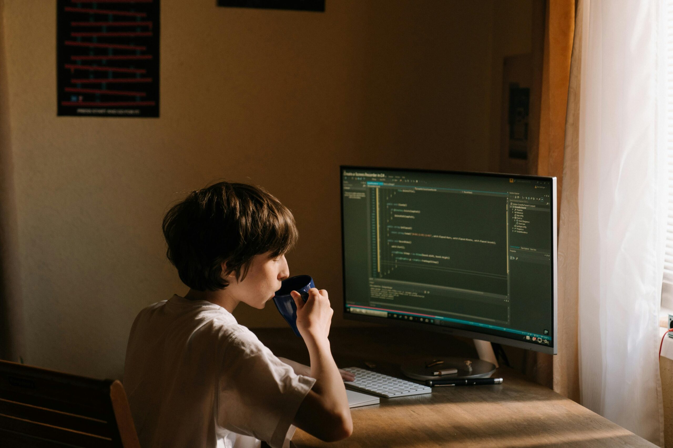A person sits at a wooden desk, sipping from a cup, while focused on a large monitor displaying programming code.